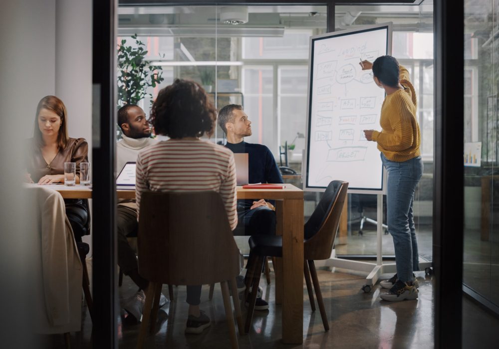 Project Manager Makes a Presentation for a Young Diverse Creative Team in Meeting Room in an Agency. Colleagues Sit Behind Conference Table and Discuss Business Development, User Interface and Design.