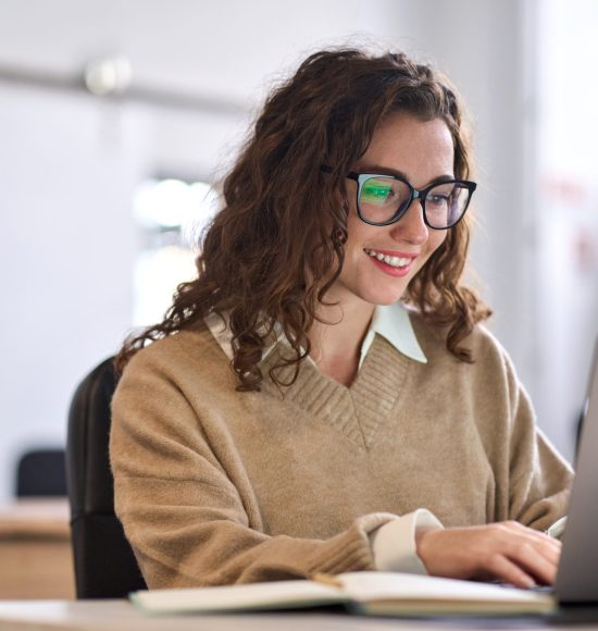 Young happy professional business woman worker employee sitting at desk working on laptop in corporate office. Smiling female student using computer technology learning online, doing web research.